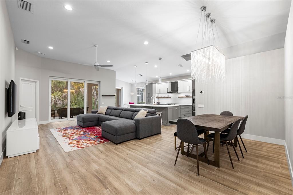 An open-concept living and dining area features wood-look tile flooring, a gray sectional, and chandelier — 8322 WHISPERING WOODS COURT, BRADENTON, FL