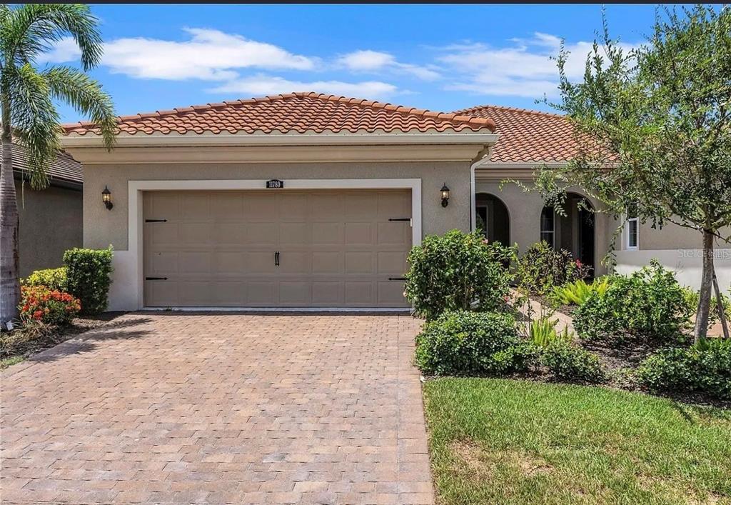 A beige house exterior features a two-car garage, a paver driveway, and a palm tree — 11780 TAPESTRY LANE, VENICE, FL