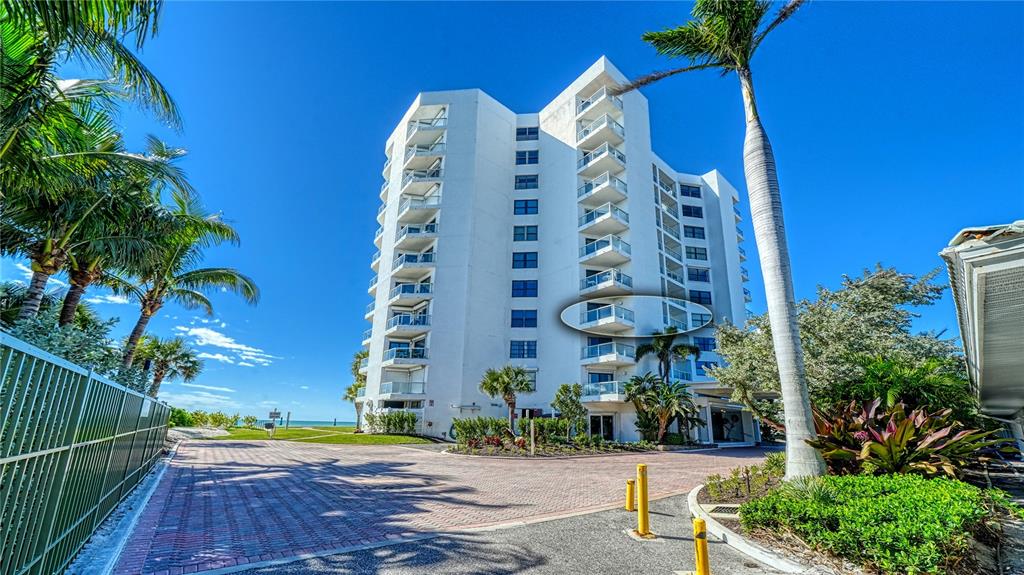 A multi-story white residential building stands near a beach with palm trees and landscaping — 1000 LONGBOAT CLUB ROAD UNIT 304, LONGBOAT KEY, FL