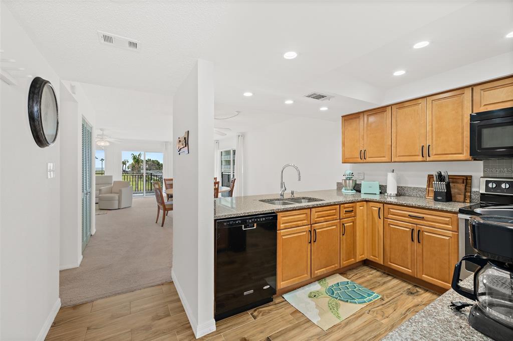 A kitchen with wood cabinets, granite countertops, and a black dishwasher opens to a living area — 1700 N ATLANTIC AVENUE UNIT 126, COCOA BEACH, FL