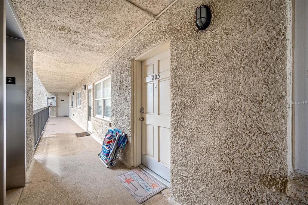 An outdoor apartment hallway features a textured stucco wall, a numbered door, and beach chairs — 19450 GULF BOULEVARD UNIT 203, INDIAN SHORES, FL