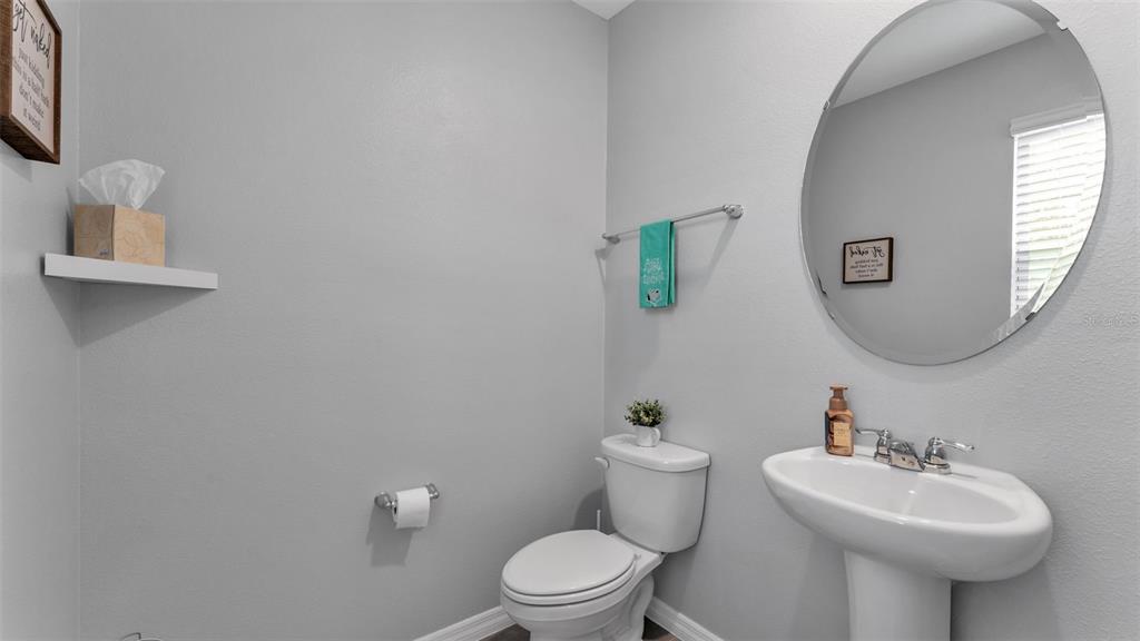 A half bathroom features a pedestal sink, round mirror, toilet, and light gray walls — 12003 HOLYSTONE STREET, WINTER GARDEN, FL