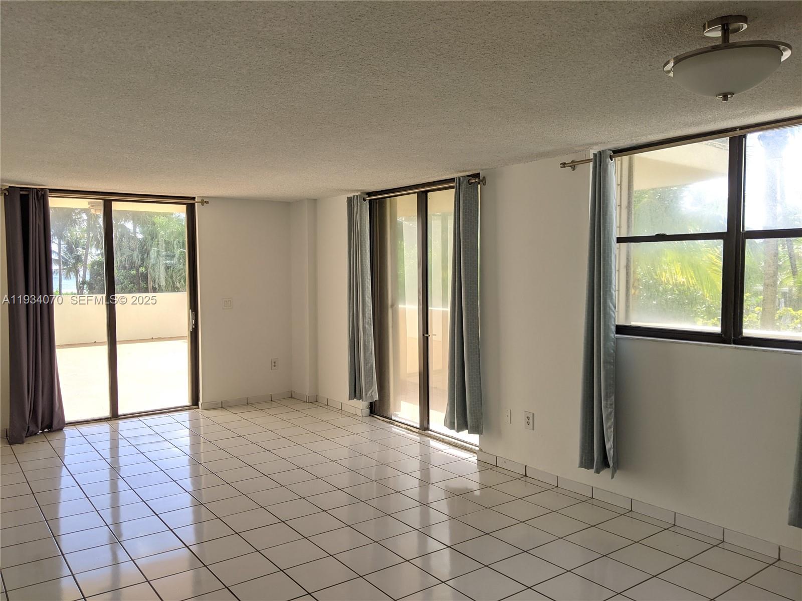 An empty room features white tile flooring, sliding glass doors, and light blue curtains — 1623 Collins Ave # 218, Miami Beach FL 33139, MIAMI BEACH, FL