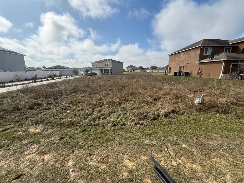 A vacant, grassy residential lot sits between two suburban houses under a cloudy blue sky — 2173 ROCK DRIVE, KISSIMMEE, FL
