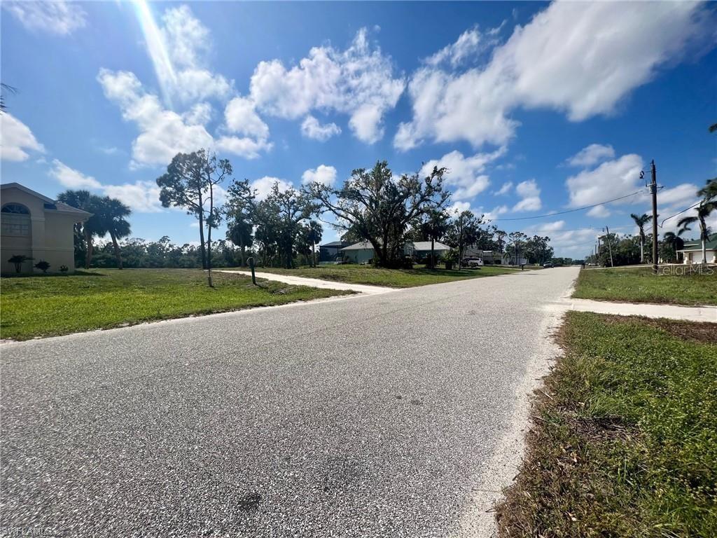 A paved residential street leads toward a grassy lot with trees under a blue sky — 5 PINE VALLEY COURT, ROTONDA WEST, FL