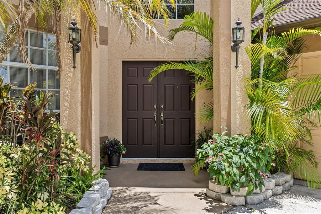 A brown double front door is framed by two pillars and lush tropical landscaping — 27221 SEA BREEZE WAY, WESLEY CHAPEL, FL