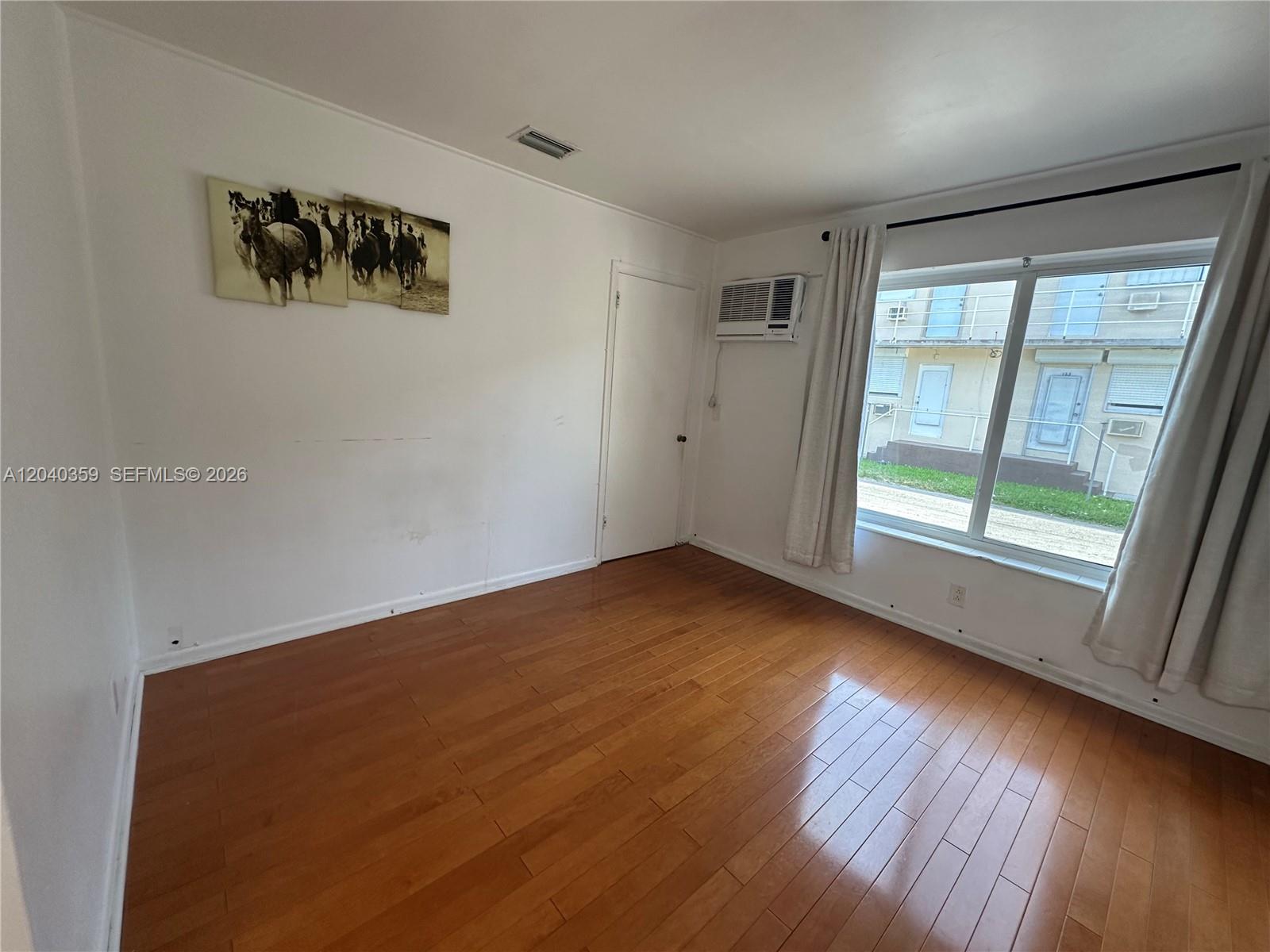 An empty bedroom features hardwood floors, white walls, a window with curtains, and an air conditioner — 819 NE 8th St # 2, Hallandale Beach FL 33009, HALLANDALE BEACH, FL