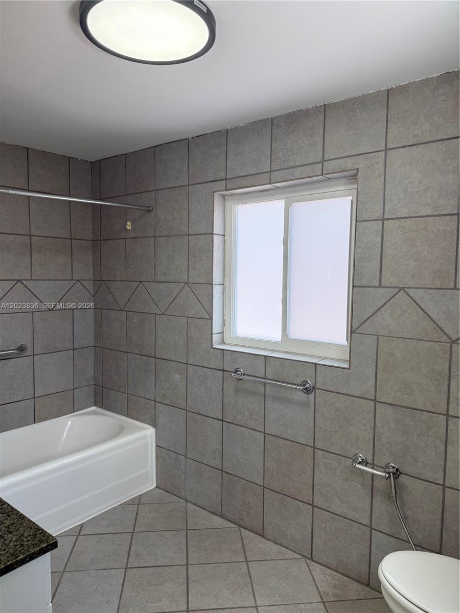 A bathroom features gray tiled walls, a white bathtub, and a window with frosted glass — 6312 Alton Rd, Miami Beach FL 33141, MIAMI BEACH, FL