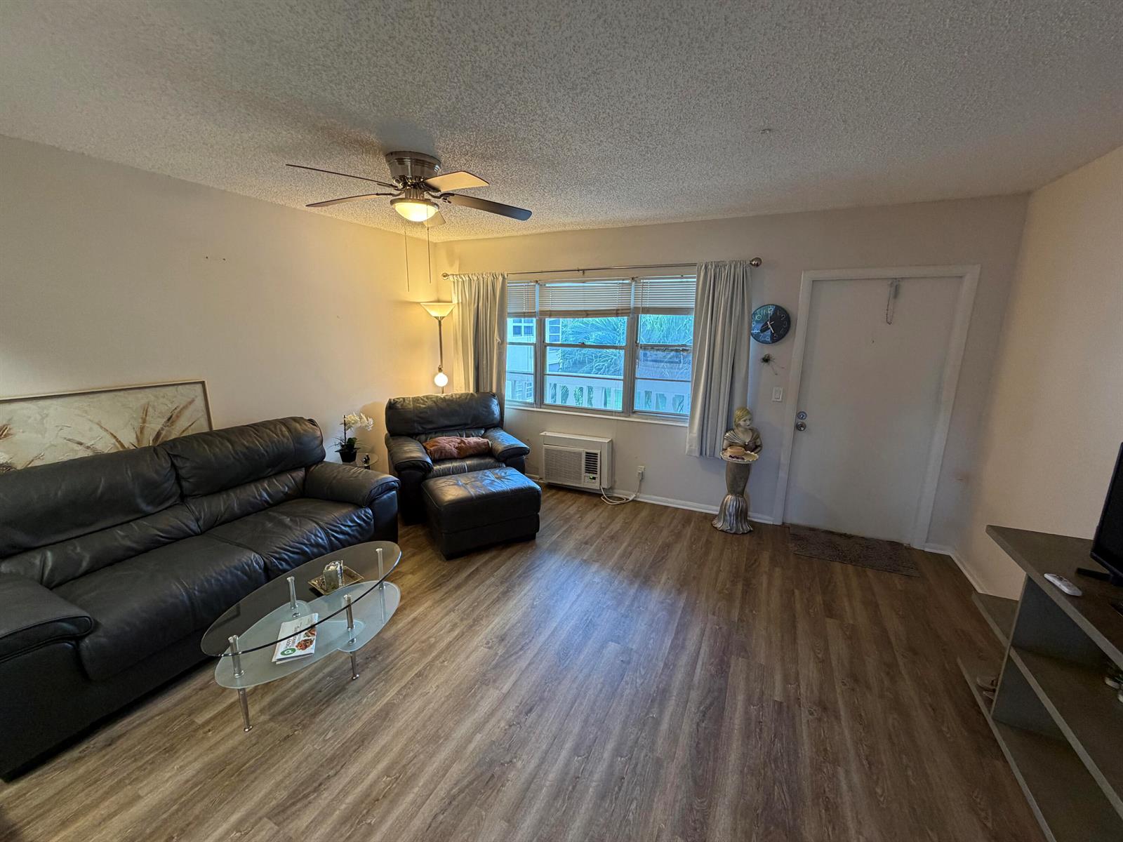 A living room features dark leather furniture, wood-look flooring, a ceiling fan, and a window — 107 Prescott E, Deerfield Beach FL 33442, DEERFIELD BEACH, FL