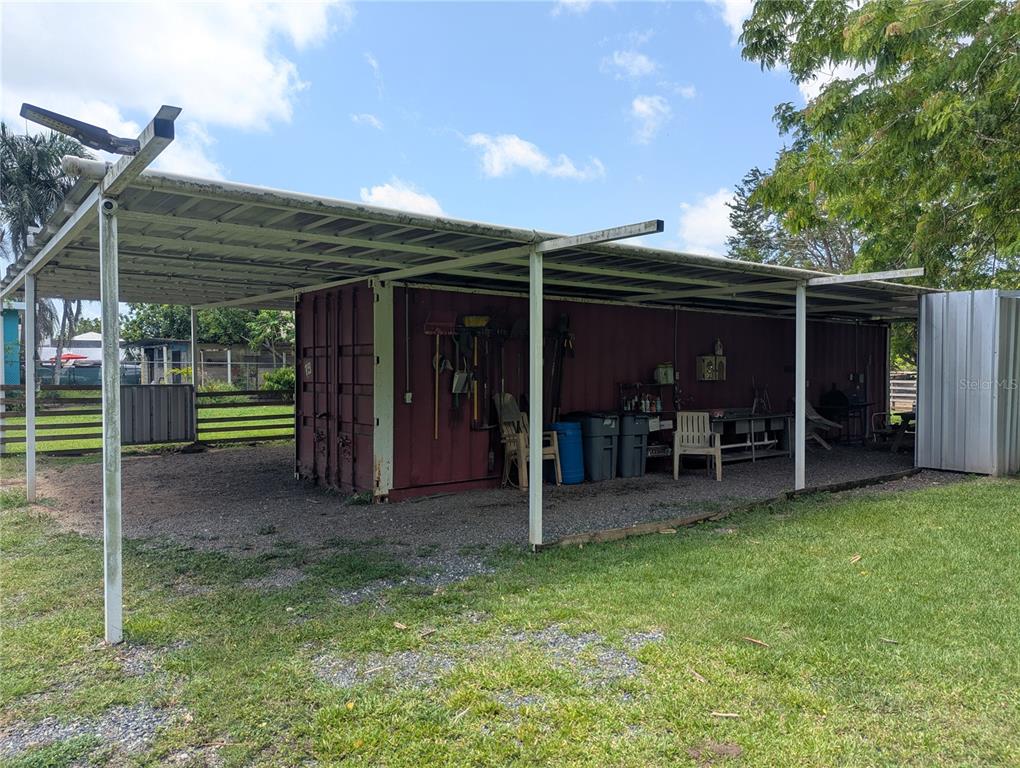 A shipping container sits under a metal carport structure on a gravel lot — BO ESPINAL, AGUADA, PR