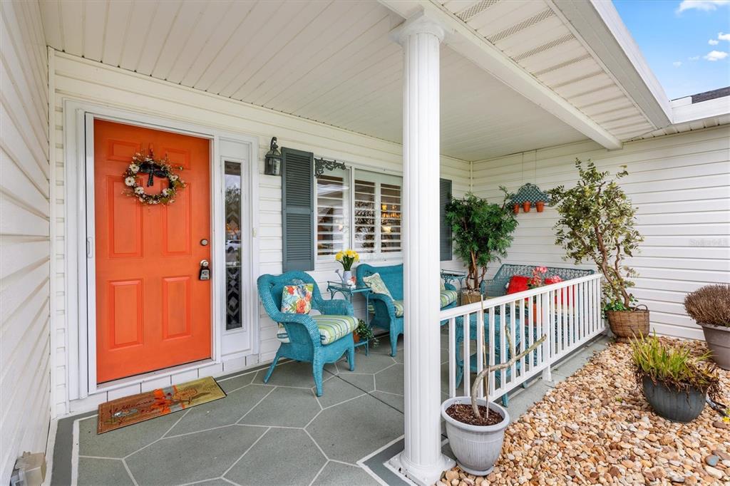 A covered front porch features a bright orange door, blue wicker furniture, and stone flooring — 17990 SE 100TH TERRACE, SUMMERFIELD, FL