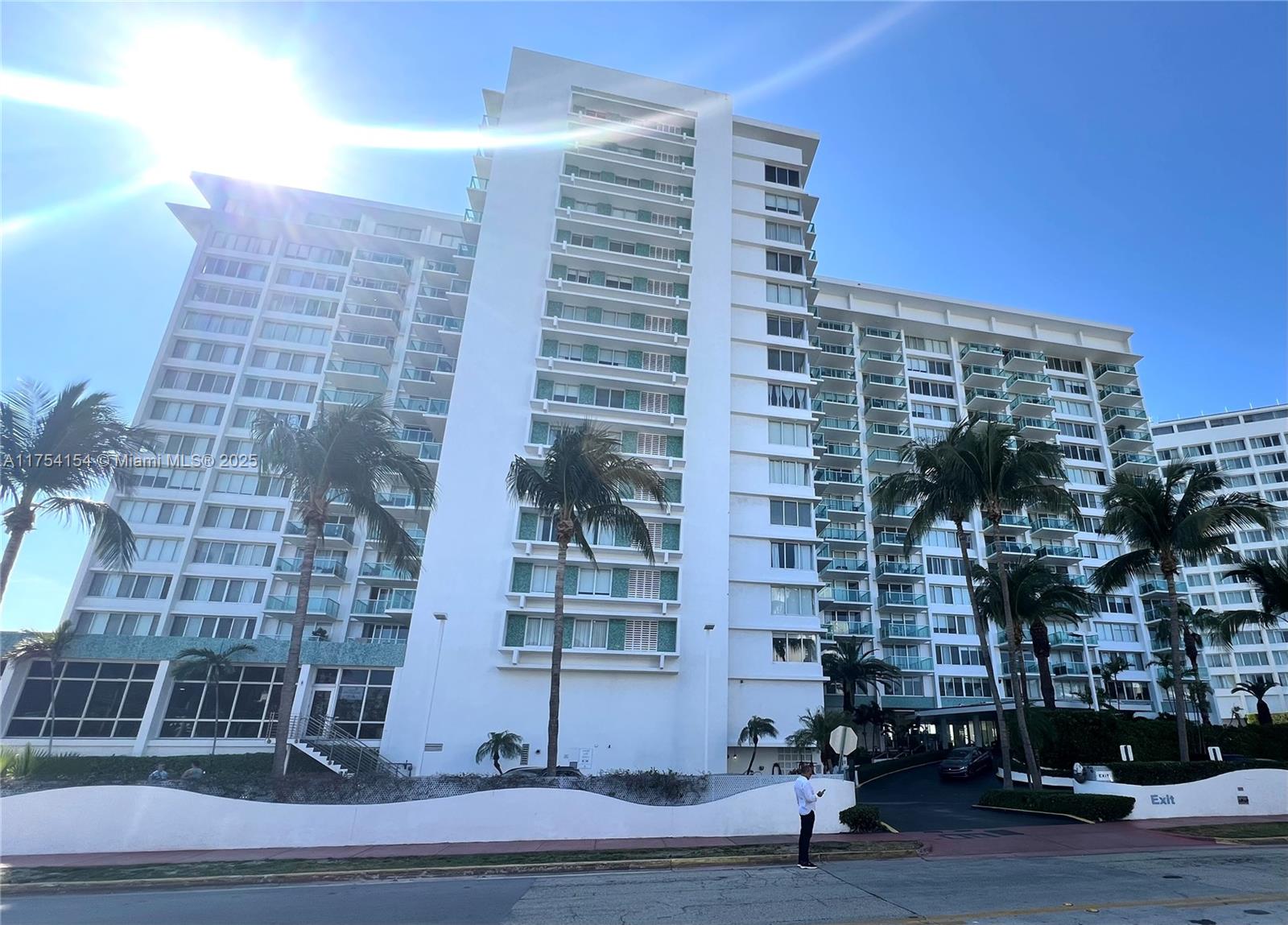A white multi-story residential building with balconies stands behind palm trees and a low wall — 1000 West Ave # 908, Miami Beach FL 33139, MIAMI BEACH, FL