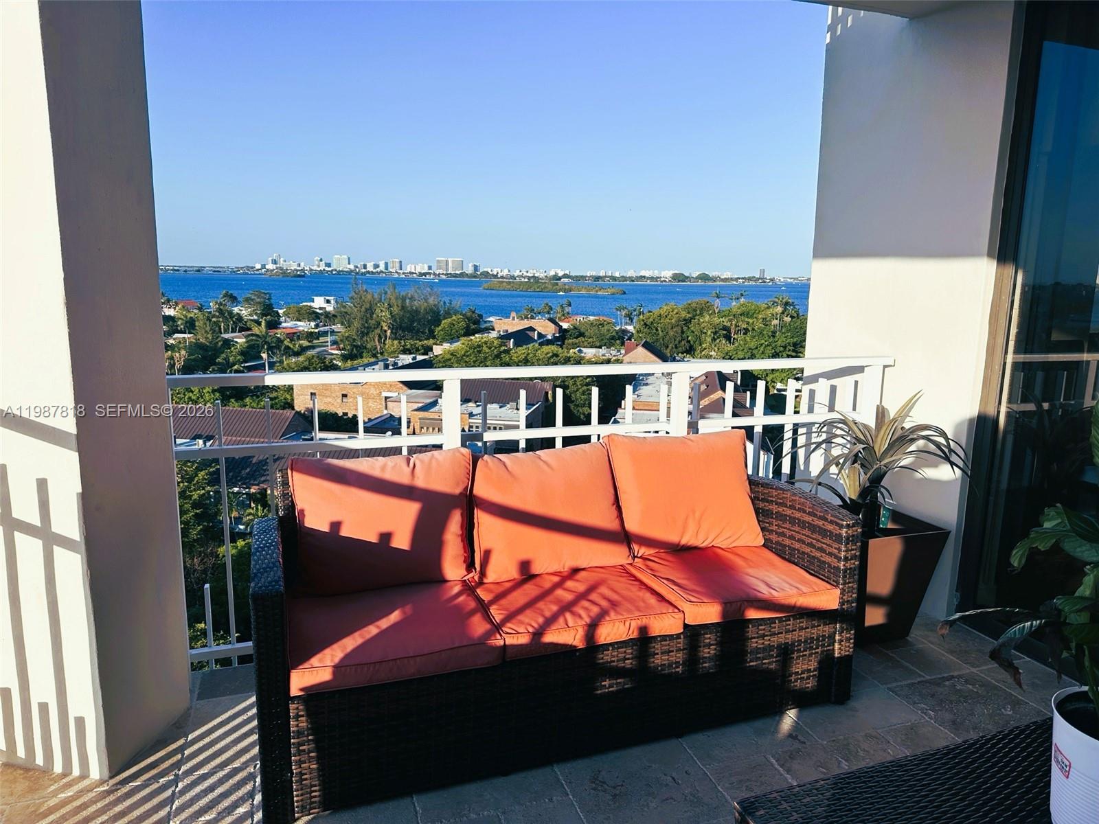 A balcony features a wicker sofa with orange cushions overlooking a bay and city skyline — 1000 Quayside Ter # 1101, Miami FL 33138, MIAMI, FL