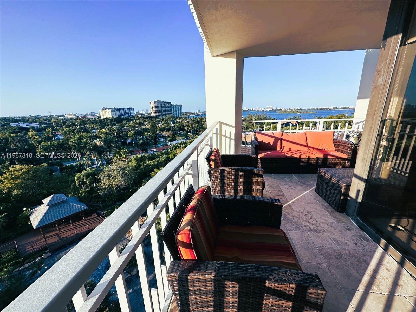 A balcony with wicker furniture overlooks a lush landscape, a gazebo, and a distant bay — 1000 Quayside Ter # 1101, Miami FL 33138, MIAMI, FL