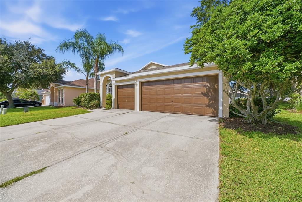 A tan house with a three-car garage and a concrete driveway under a blue sky — 5553 BRADDOCK DRIVE, ZEPHYRHILLS, FL