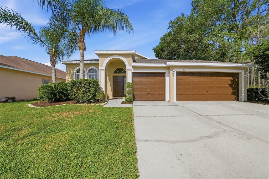 A single-story house with a tan stucco exterior, a three-car garage, and a palm tree — 5553 BRADDOCK DRIVE, ZEPHYRHILLS, FL