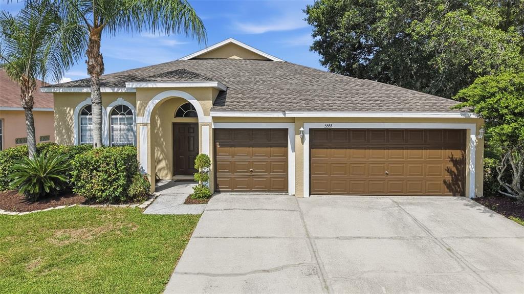A single-story house with a tan stucco exterior, brown garage doors, and a palm tree — 5553 BRADDOCK DRIVE, ZEPHYRHILLS, FL