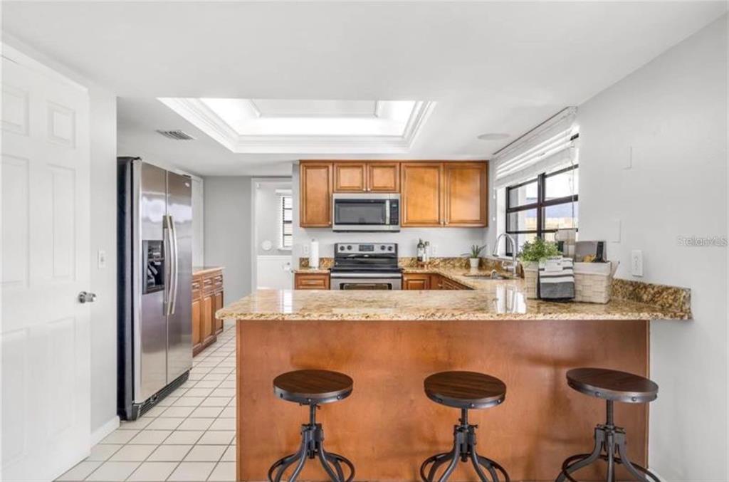 A kitchen features wood cabinets, granite countertops, a breakfast bar with stools, and tile flooring — 16208 PEBBLEBROOK DRIVE, TAMPA, FL