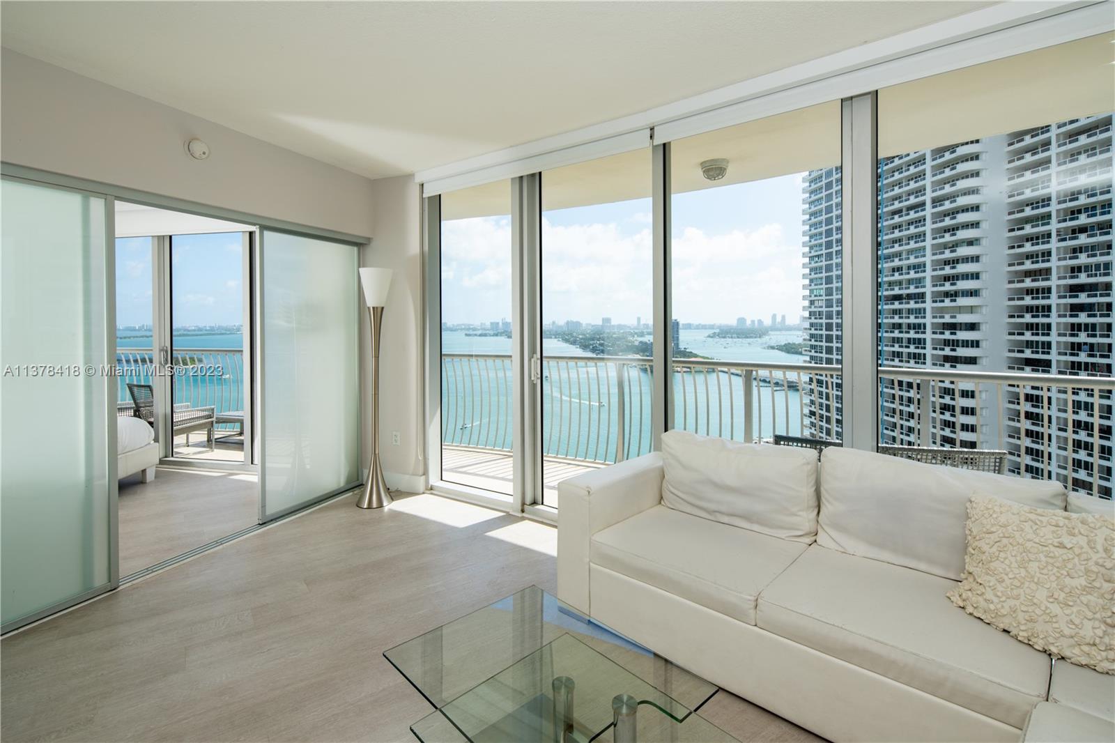 A living room features a white sofa, glass coffee table, and floor-to-ceiling windows overlooking water — 1750 N Bayshore Dr # 2602, Miami FL 33132, MIAMI, FL