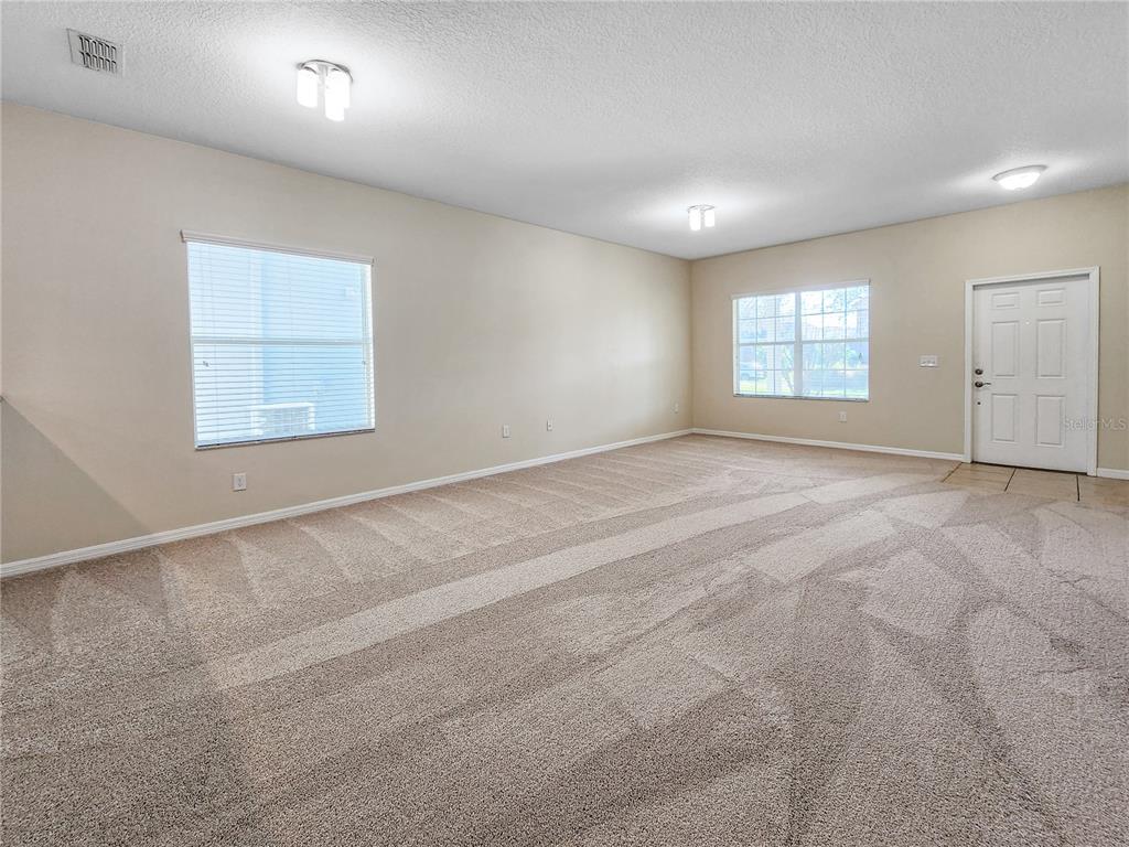 An empty living room features beige walls, neutral carpeting, two windows, and a white door — 6843 POINT HANCOCK DRIVE, WINTER GARDEN, FL