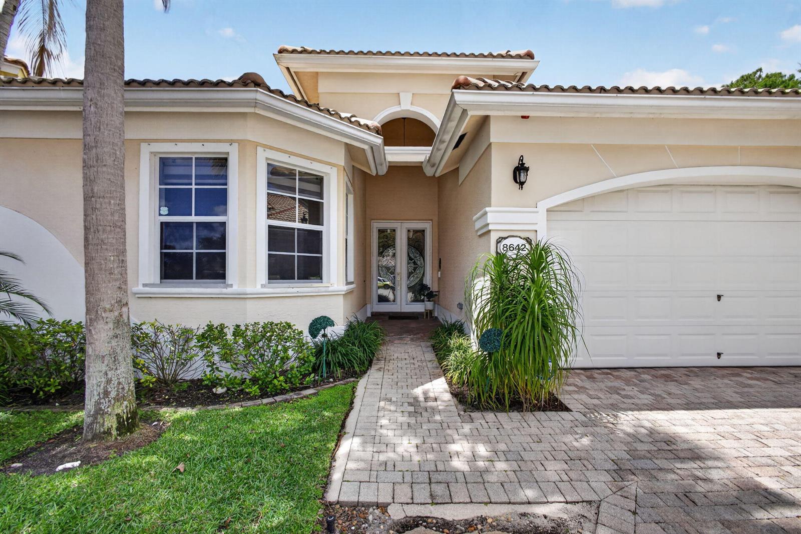 The exterior of a beige house features a paved walkway, a garage, and a front entrance — 8642 Plum Cay, Royal Palm Beach FL 33411, ROYAL PALM BEACH, FL