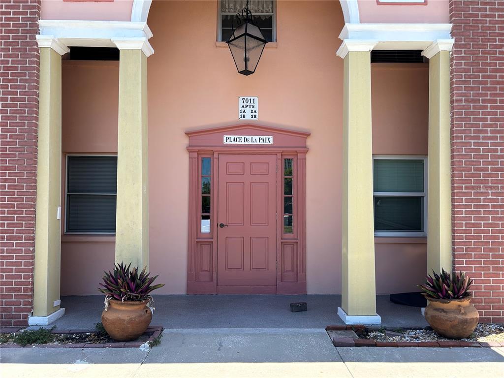 The exterior entrance of a building features a pink door, two columns, and potted plants — 7011 PLACE DE LA PAIX UNIT 1A, ST PETERSBURG, FL