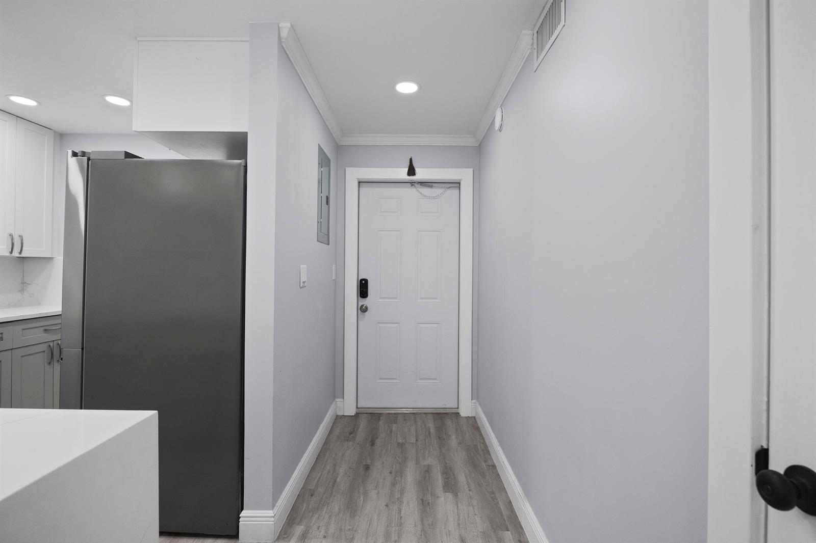 A hallway with light gray walls, wood-look flooring, and a white door leads to an entrance — 9856 Marina Boulevard # 1314, Boca Raton FL 33428, BOCA RATON, FL