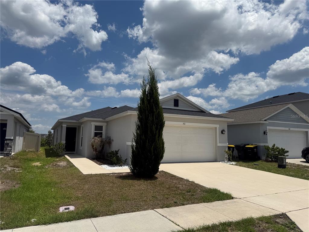 A single-story suburban house with a white garage, light-colored stucco exterior, and a tall evergreen — 711 SCRUB OAK HAMMOCK ROAD, DAVENPORT, FL