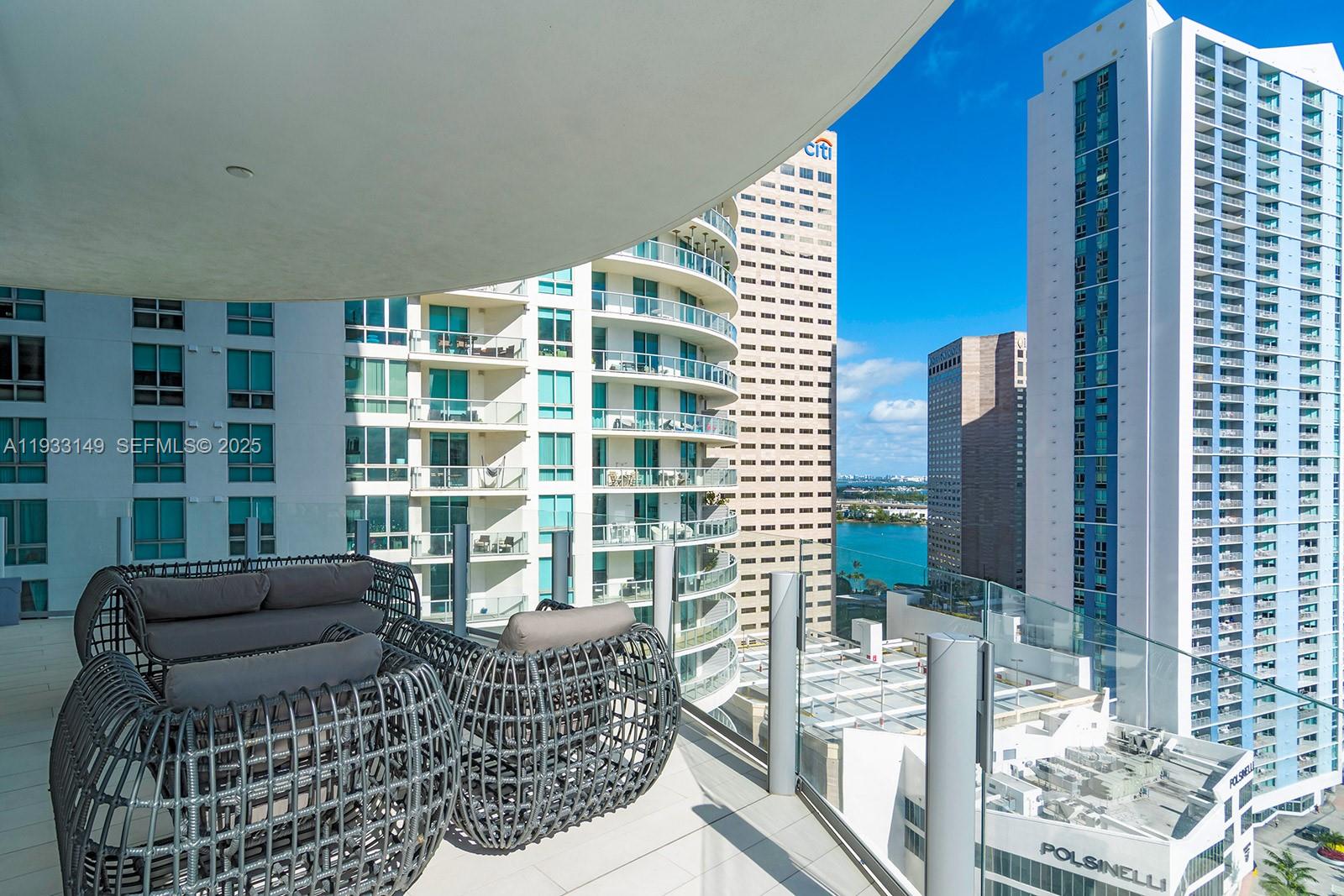 A high-rise balcony features modern wicker furniture overlooking a city skyline and water — 300 Biscayne Blvd Way # 1901, Miami FL 33131, MIAMI, FL