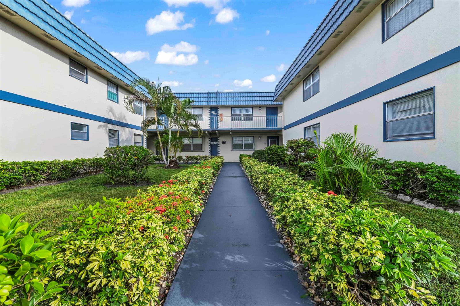 A paved walkway leads between two apartment buildings with blue roofs and landscaped garden beds — 213 Brittany, Delray Beach FL 33446, DELRAY BEACH, FL