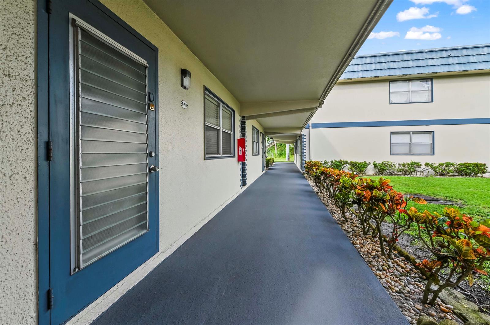 An exterior walkway leads past a blue door with a louvered window and landscaping — 213 Brittany, Delray Beach FL 33446, DELRAY BEACH, FL