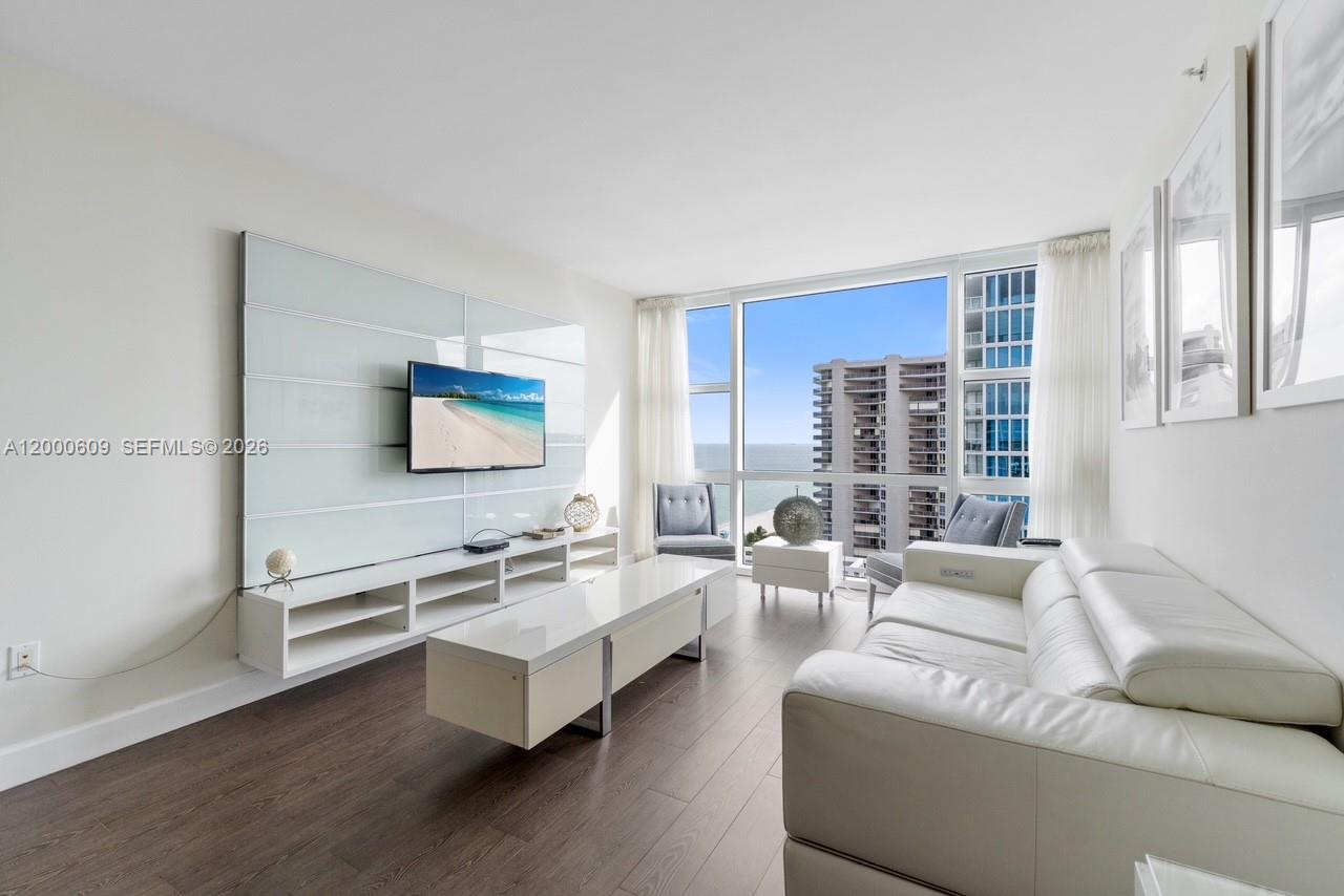 A modern living room features a white media console, white leather sofa, and floor-to-ceiling windows — 6801 Collins Ave, Miami Beach FL 33141, MIAMI BEACH, FL
