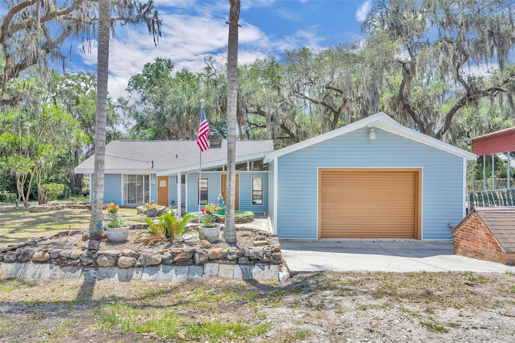 A light blue single-story house features a garage, a stone retaining wall, and mature trees — 13450 SE 105TH TERRACE, OCKLAWAHA, FL