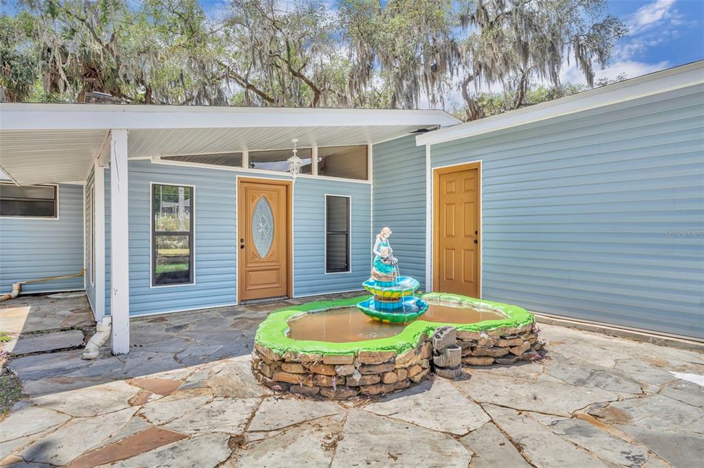 A blue-sided house exterior features a stone patio, a tiered fountain, and two wooden doors — 13450 SE 105TH TERRACE, OCKLAWAHA, FL