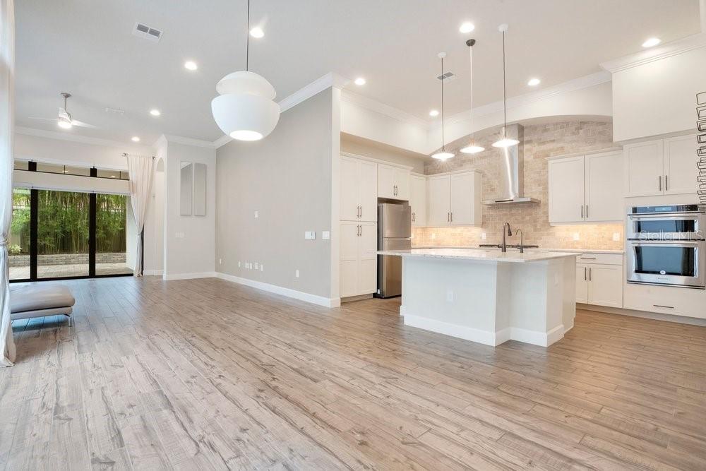 A modern kitchen with white cabinetry, a center island, and wood-look flooring opens to a living area — 8394 VIA VITTORIA WAY, ORLANDO, FL