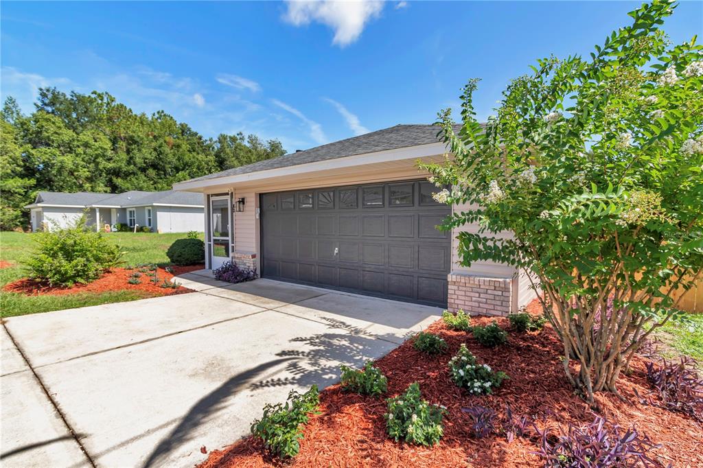 A single-story house exterior features a gray garage door, a concrete driveway, and landscaped mulch beds — 63 PECAN PASS, OCALA, FL