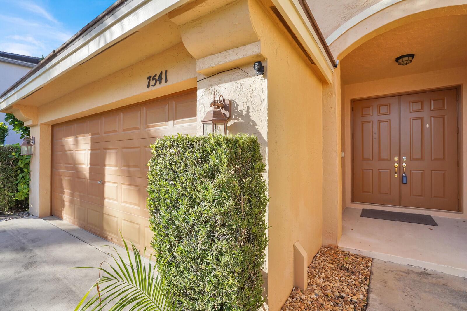 A tan house exterior features a double garage, a brown front door, and a hedge — 7541 Downwinds Lane, Lake Worth FL 33467, LAKE WORTH, FL