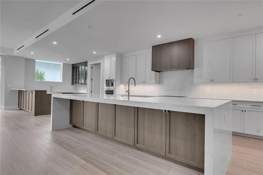 A modern kitchen features a large white waterfall island, wood cabinetry, and light tile flooring — 70 AVENIDA VENECCIA UNIT 204, SARASOTA, FL