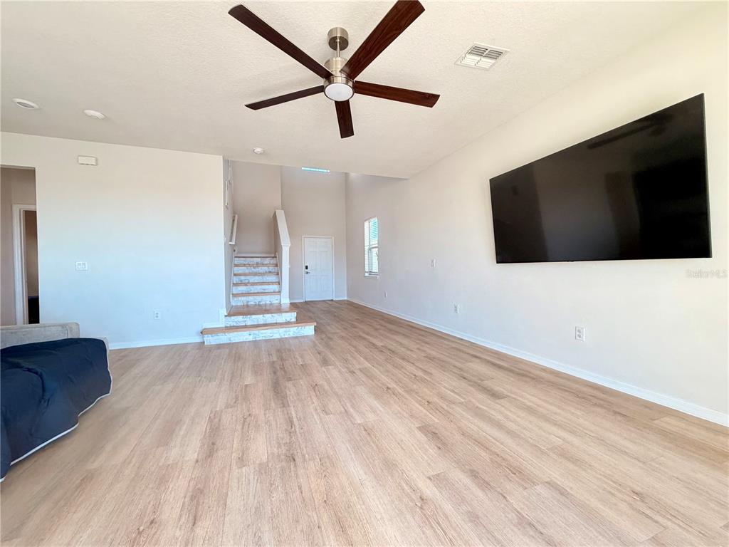 A living room features light wood-look flooring, a ceiling fan, and a large wall-mounted television — 11415 GREEN HARVEST DRIVE, RIVERVIEW, FL