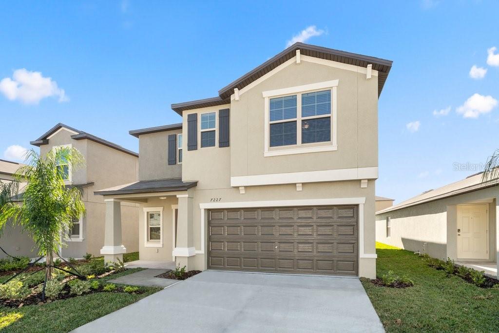 A two-story suburban house features a tan stucco exterior, a brown garage door, and landscaping — 7227 RONNIE GARDENS COURT, TAMPA, FL