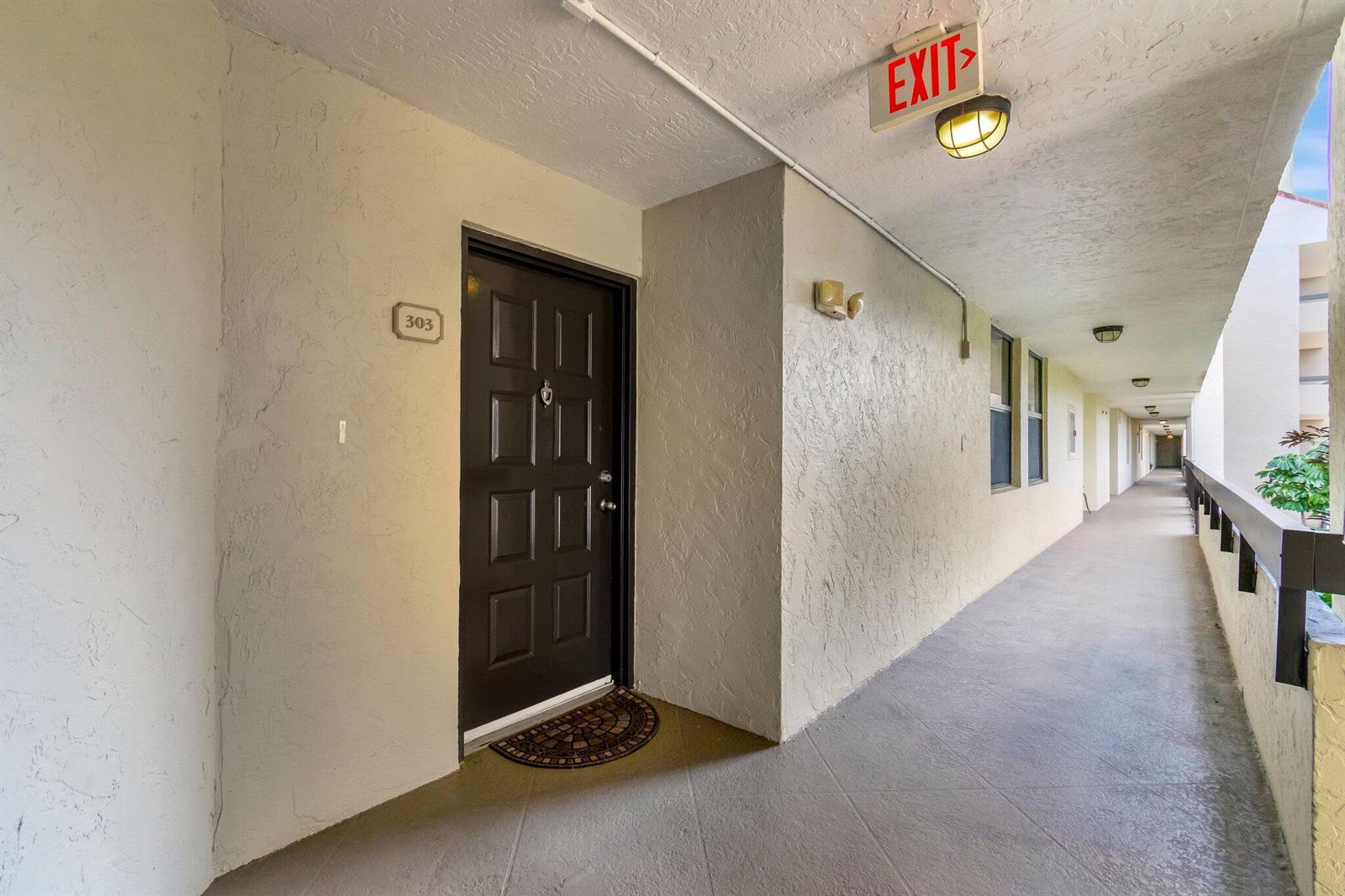 An exterior apartment hallway features a dark brown door labeled 303 and an overhead exit sign — 1915 Lavers Circle # 303, Delray Beach FL 33444, DELRAY BEACH, FL