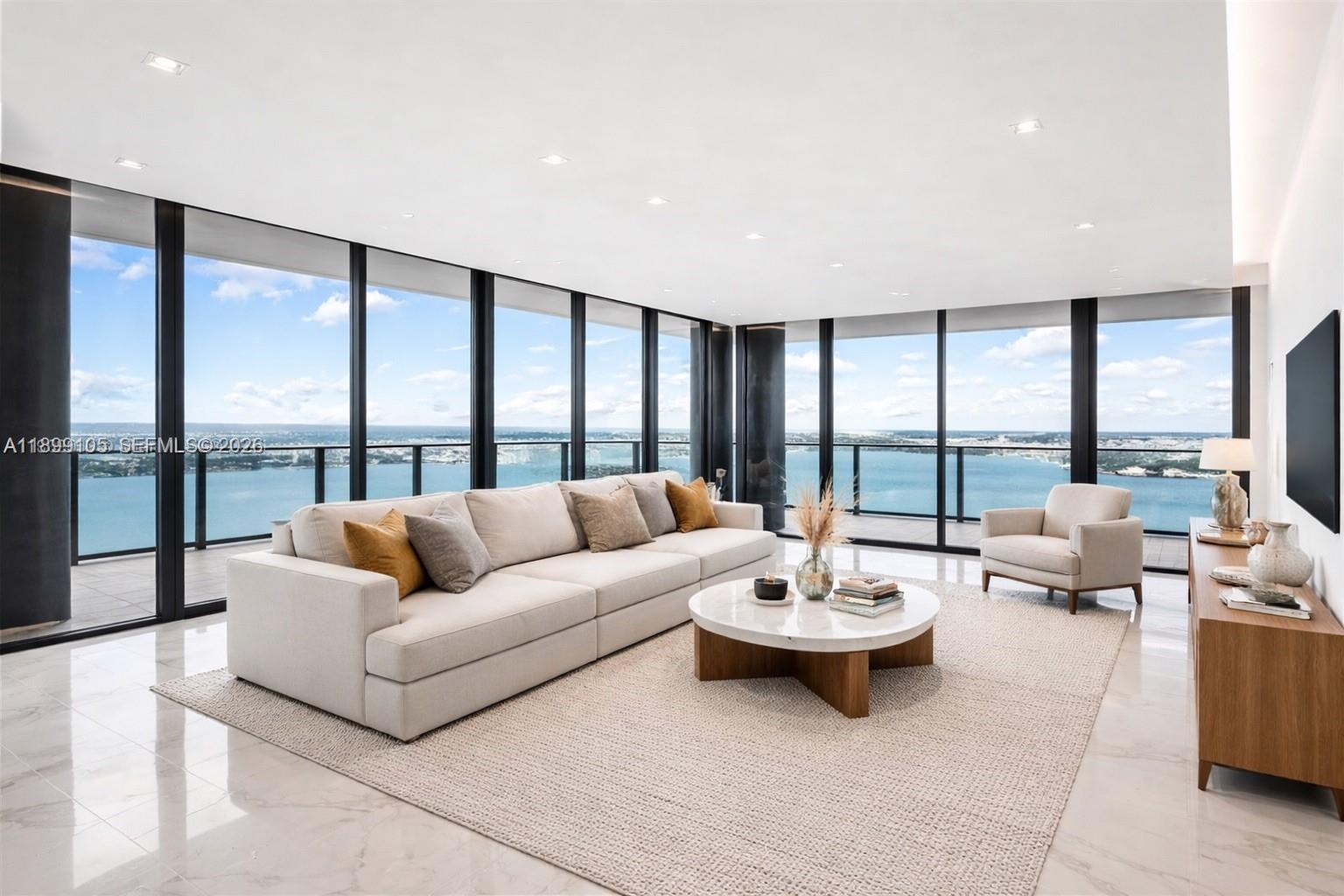 A modern living room features a large beige sofa, marble floors, and floor-to-ceiling windows overlooking water — 700 NE 24th St # 5401, Miami FL 33137, MIAMI, FL