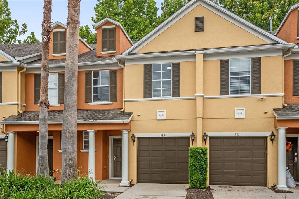 A two-story townhouse exterior with a tan stucco facade, brown garage doors, and palm trees — 815 ASSEMBLY COURT, KISSIMMEE, FL
