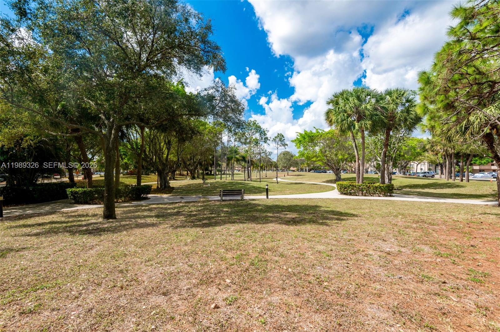 A paved walkway winds through a grassy park area with mature trees and a bench — 6301 N Fall Circle Dr # 213, Lauderhill FL 33319, LAUDERHILL, FL