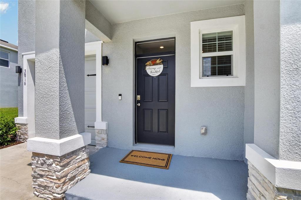 A gray front door with a wreath and a welcome mat sits under a covered porch — 3816 SUNFLOWER FIELD PLACE, PLANT CITY, FL
