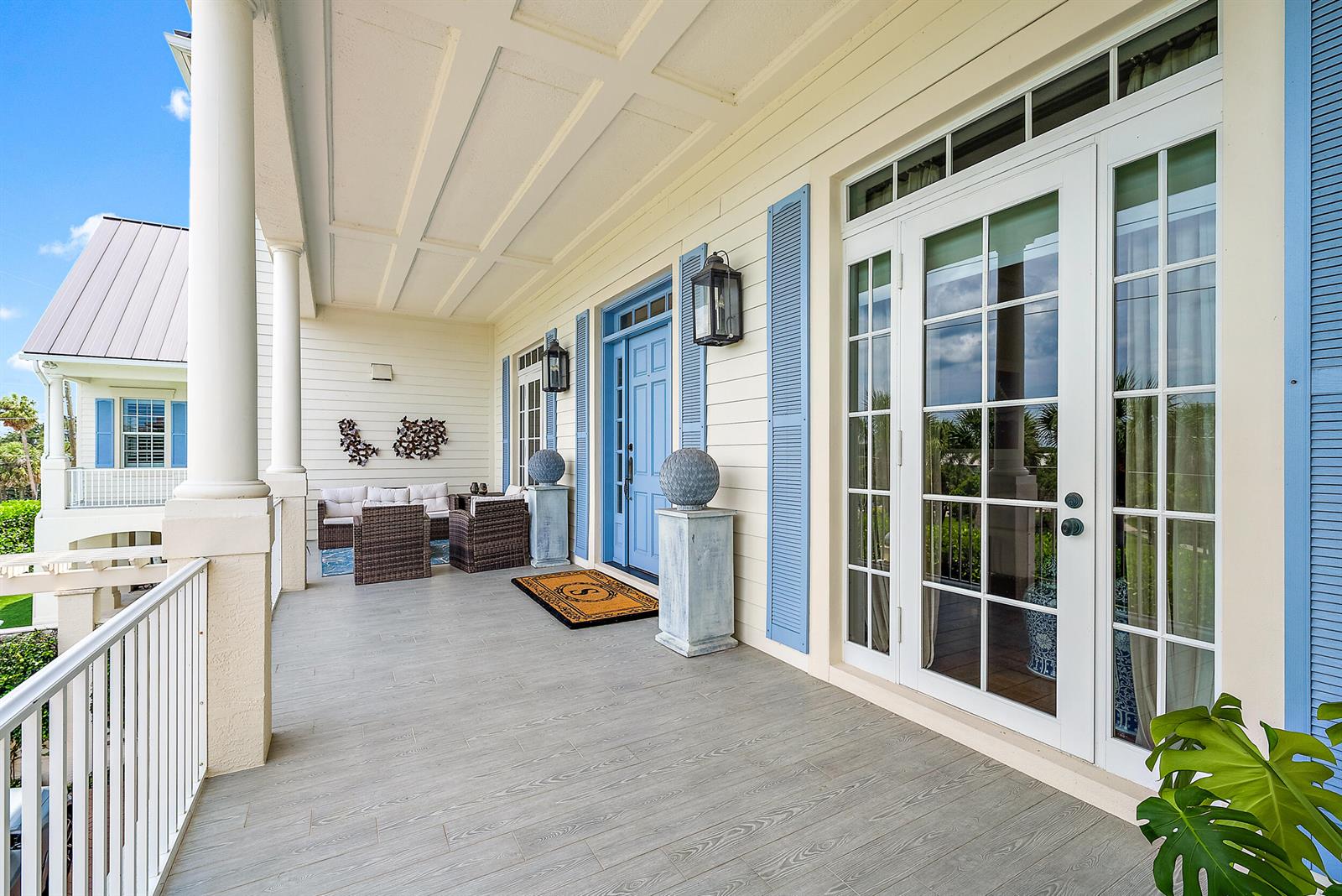 A covered porch features light wood-look flooring, blue shutters, a blue front door, and wicker furniture — 410 Old Towne Lane, Juno Beach FL 33408, JUNO BEACH, FL