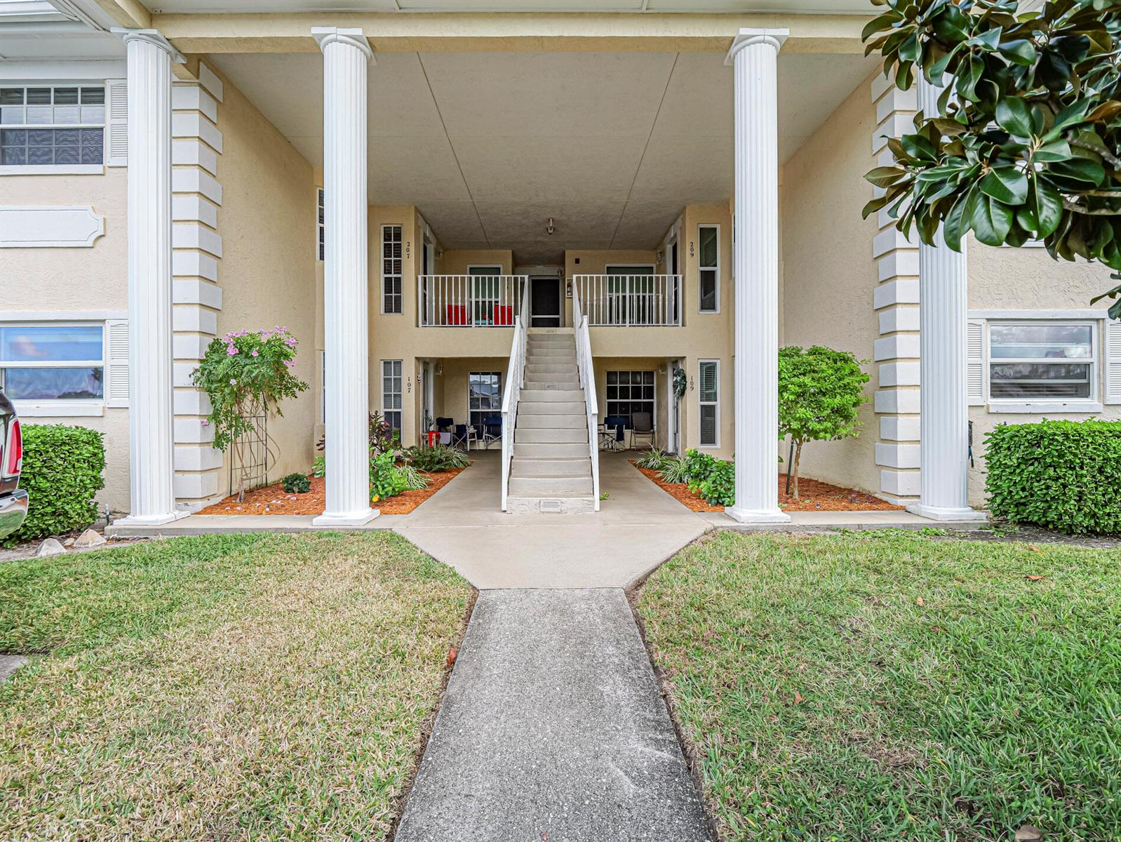 A concrete walkway leads to a two-story apartment building entrance with white columns and stairs — 700 Lake Orchid Circle # 209, Vero Beach FL 32962, VERO BEACH, FL