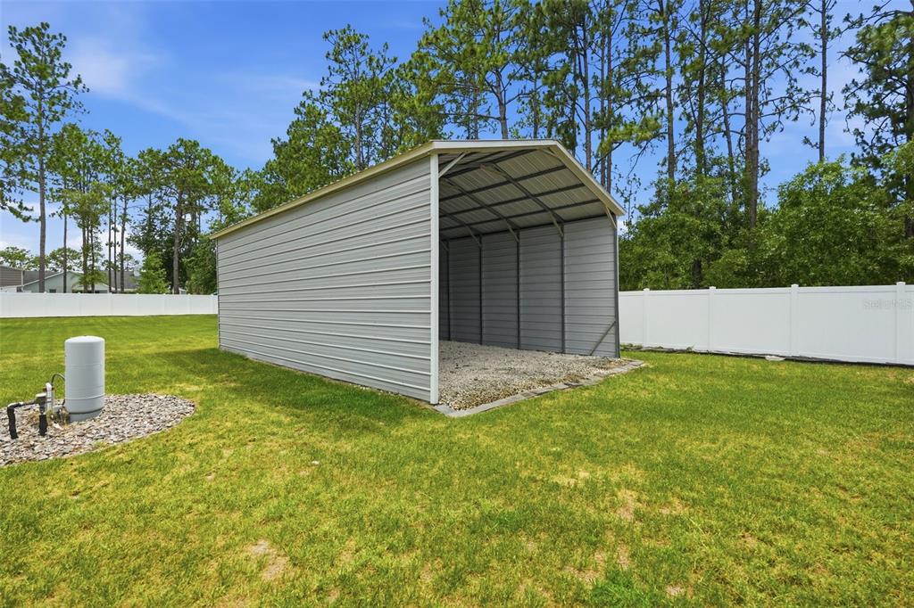 A metal carport with a gravel floor sits in a grassy backyard near a fence — 4743 JUMPER LOOP, SPRING HILL, FL