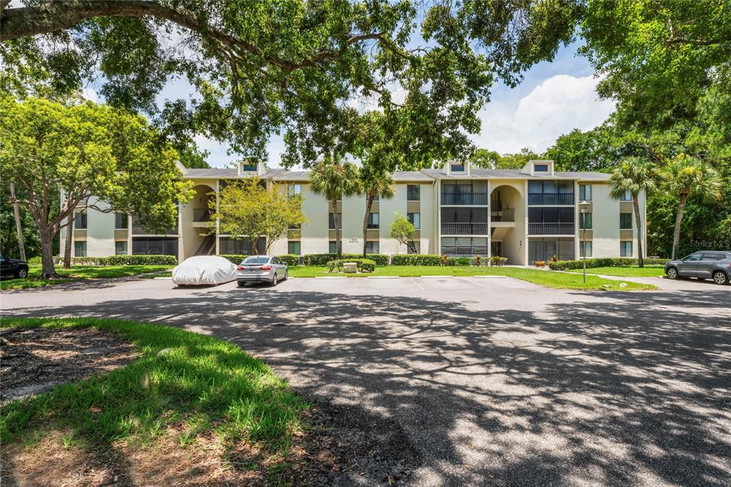 A three-story apartment building with a parking lot and mature trees in the foreground — 3232 LAKE PINE WAY UNIT H2, TARPON SPRINGS, FL