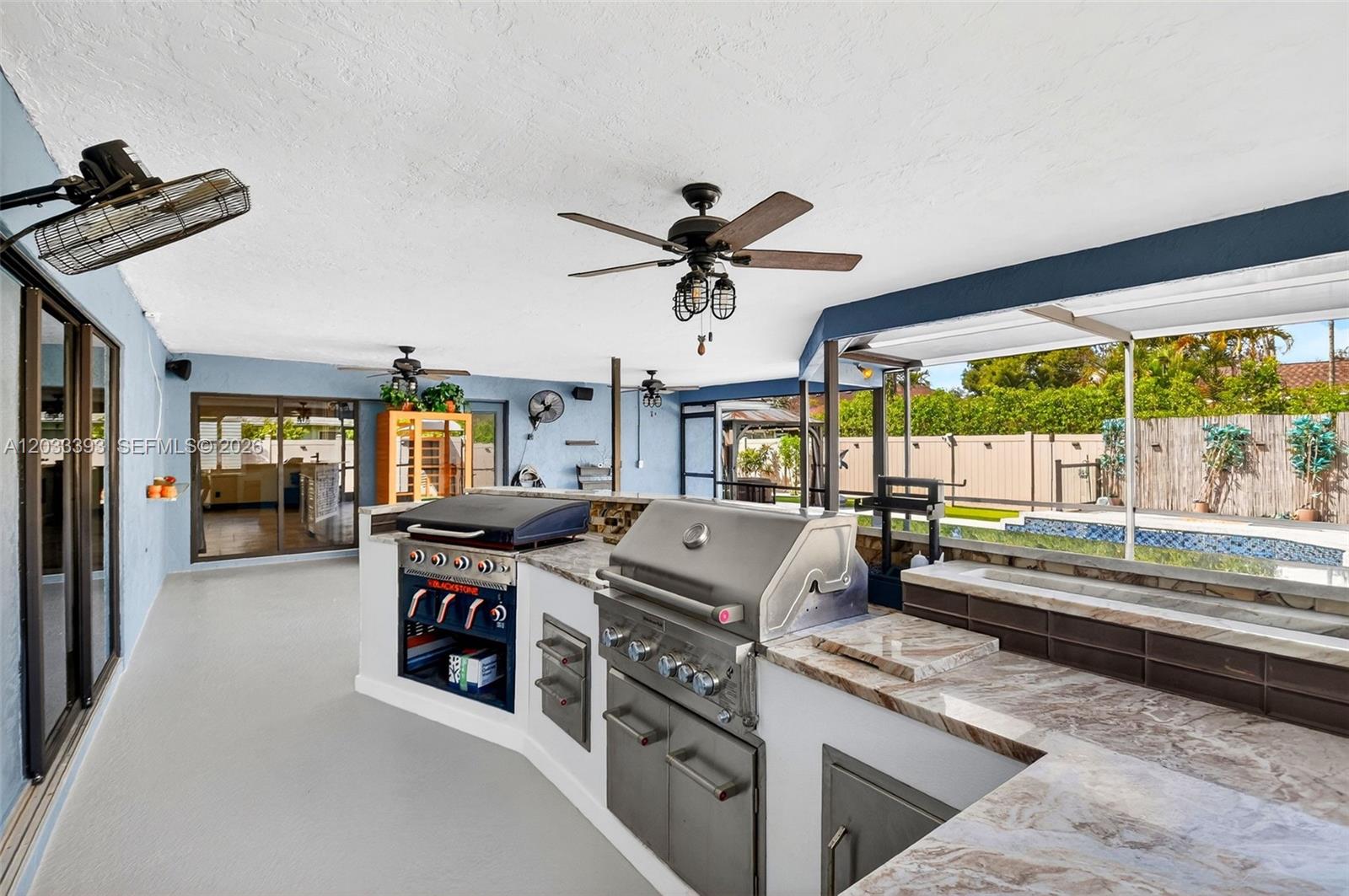An outdoor kitchen features a stainless steel grill, a griddle, and marble countertops under a patio — 13808 Geranium Pl, Wellington FL 33414, WELLINGTON, FL